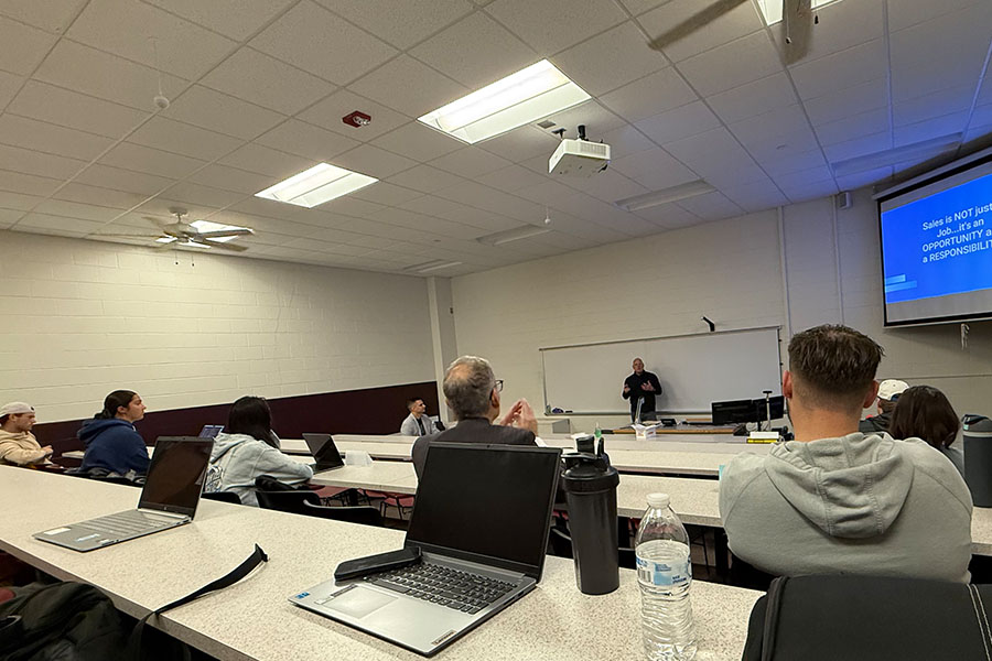 Ron Alvesteffer stands in the front of a classroom wearing a long-sleeve black shirt. Behind him is a white board and above him is a display screen with his presentation. Students sit at long tables in the foreground and watch him as he speaks.