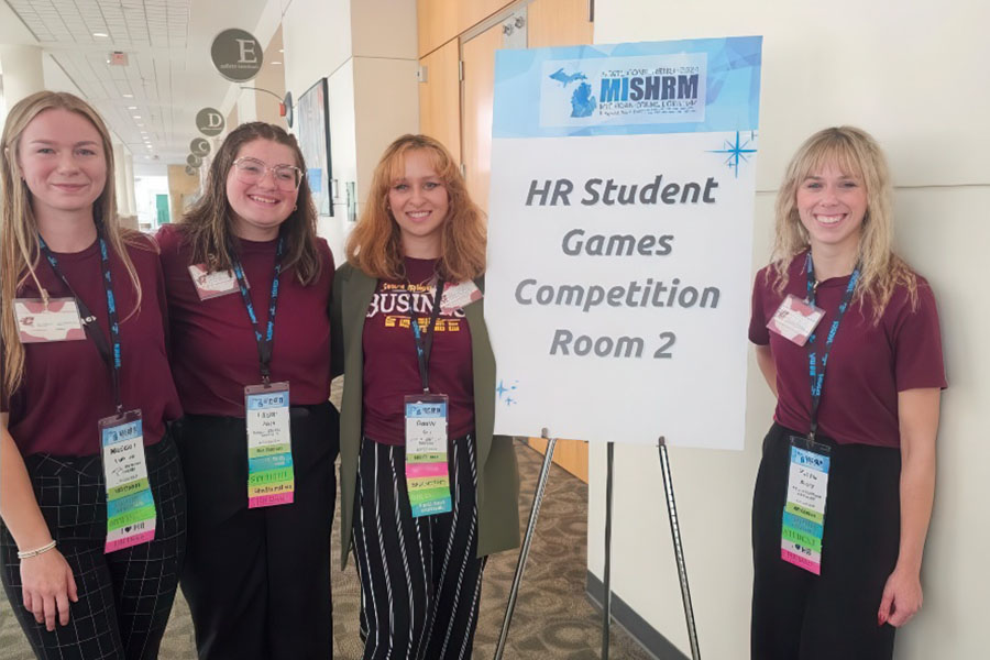 Four SHRM student members stand with matching maroon shirts and event lanyards around a sign that reads
