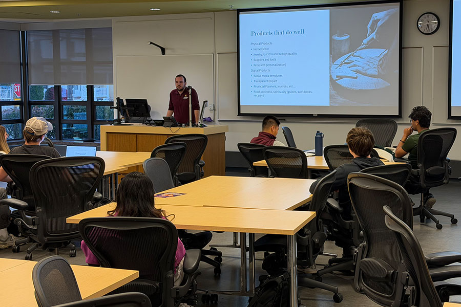 Daniel Lahey stands in front of students in the 150G classroom. He is wearing a maroon poly shirt. Students sit at tables throughout the room and face him. Behind him is a pull-down screen with a presentation slide projected on it.