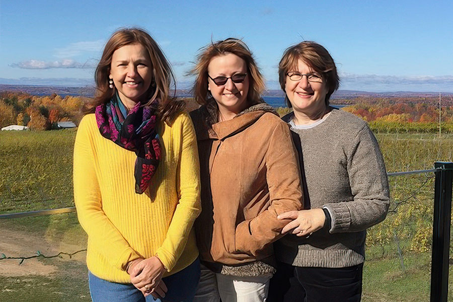 Susan Carrington appears on the left in a yellow shirt and jeans, Sarah Opperman stands in the middle in a brown jacket and white pants, and Jan Hewlett stands on the right with a tan sweater.