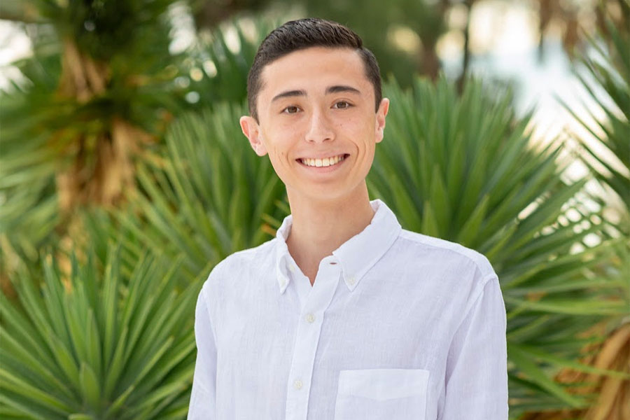 Travis Tompkins poses in front of tropical ferns and palm trees. He's wearing a white button down shirt. He has black hair that is gelled and brushed back. He has a large wide smile.
