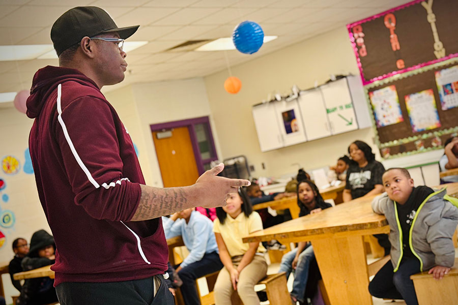 Tupac Holmes wears a maroon zip up sweatshirt with a white racing stripe down the arm. He has on a black ball cap and glasses. He's gesturing and you can see script from a tattoo on his arm. He's facing a classroom full of kids who look at him intently.