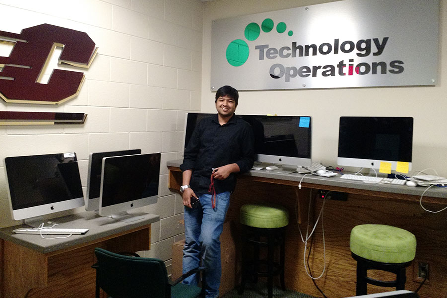 Vivek Kovvuru appears in an old photograph in an OIT office as an undergrad. He's wearing jeans and a black shirt. There are green stools and Mac computers on tables. A sign on the wall says 