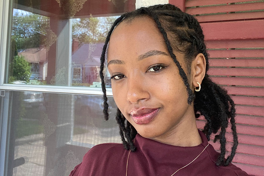 900x600_Article_Inset_Yvette Yvette stands in front of a window with a rust colored shutter. She's wearing a high neck maroon shirt with a necklace. She has dreads that are pulled back and is wearing gold hoop earrings.