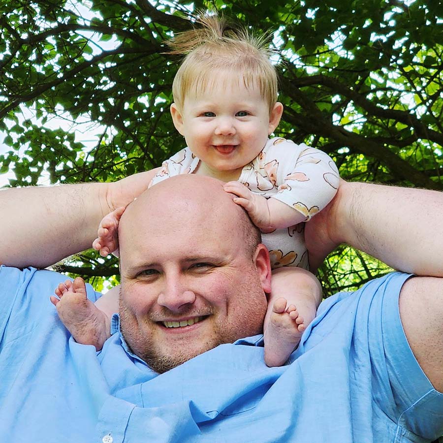 Winston Larson wears a blue button down and has grins as he holds his 9-month-old daughter on his shoulders. His daughter has a top pony tail with hair sticking up around it, fat cheeks and squinty blue eyes.
