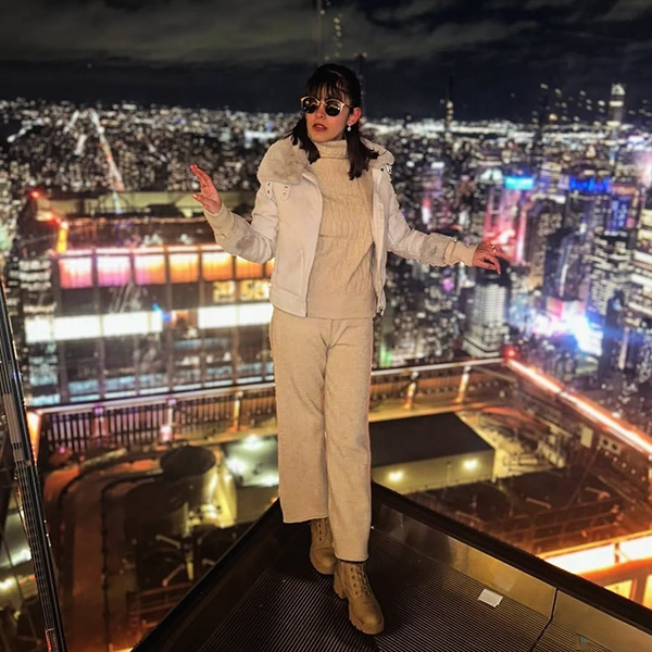 Article_Inset_600x600_Aimen_Cityscape Aimen Khan stands on a platform overlooking a brightly-lit cityscape. She's wearing a tan sweater, tan ankle length pants, light brown boots with a chunky heel, and a white cream jacket with a fur color. She has black sunglasses.