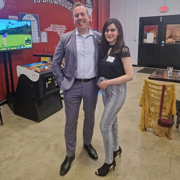 Article_Inset_600x600_Aimen_MacKinnon Aimen Khan stands with CMU President Neil MacKinnon. She wears a black short-sleeve shirt, black plaid pants and black heels. Pres. MacKinnon wears a gray suit. They are in a brewery with arcade games and casks in the background.