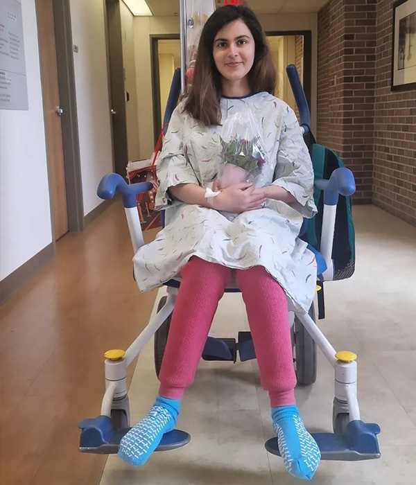 Article_Inset_600x700_Aimen_Hospital Aimen Khan sits in a wheelchair in a hospital corridor. She has long dark brown hair and smiles slightly. She wears a hospital gown over pink pants and wears blue hospital grippy socks. She holds a plant in her lap.
