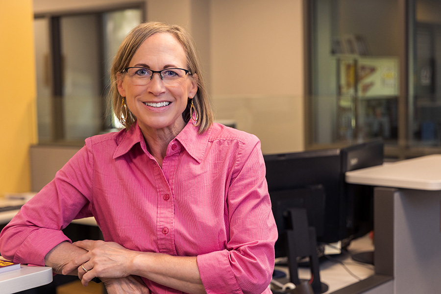 Karen Arthur stands at a desk in the Business Student Services office. She has chin length blond hair, black rimmed glassed, a pink button down shirt and dangly silver earrings.
