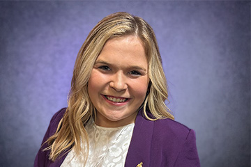 Portrait of Haylei Drope wearing a purple blazer emblazoned with a pin. She's smiling, looking into the camera, her blue eyes matching brilliantly with the gray-blue background.