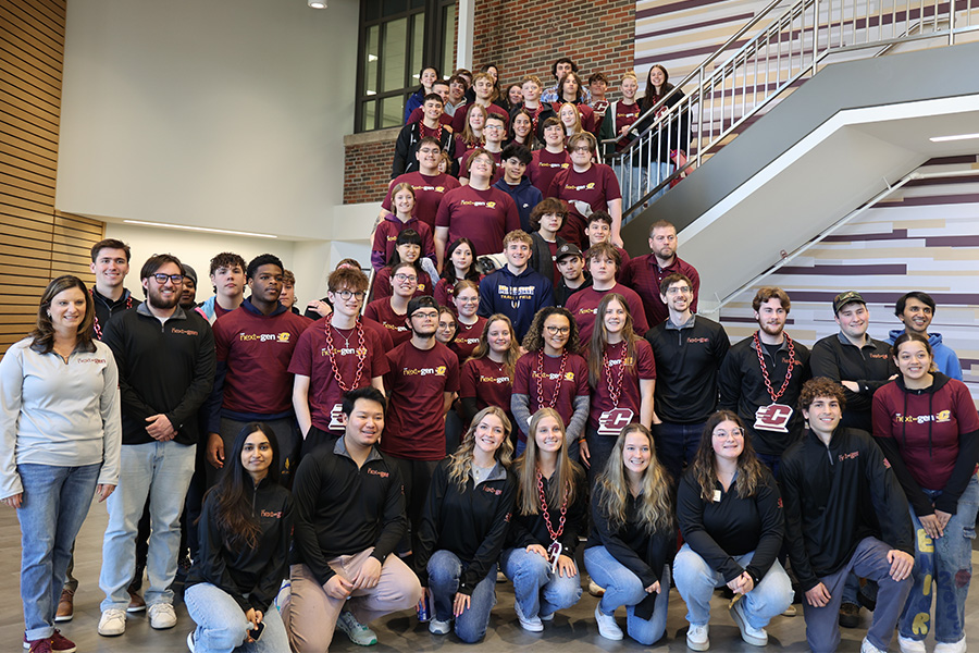 A large group of high school students, faculty members and teachers in black and maroon shirts pose on the stairs in the Grawn Atrium.