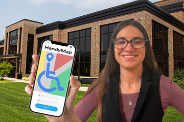 Jade Walleman stands in front of Grawn hall wearing a mauve shirt and black vest, while a hand with the HandyMap app on a cell phone screen appears from off side.
