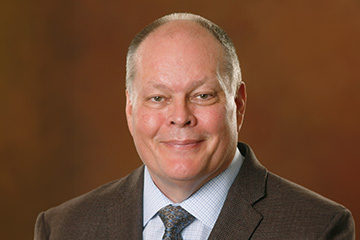 Professional Headshot of Karl Smart. He's wearing a brown suit in front of a brown backdrop.