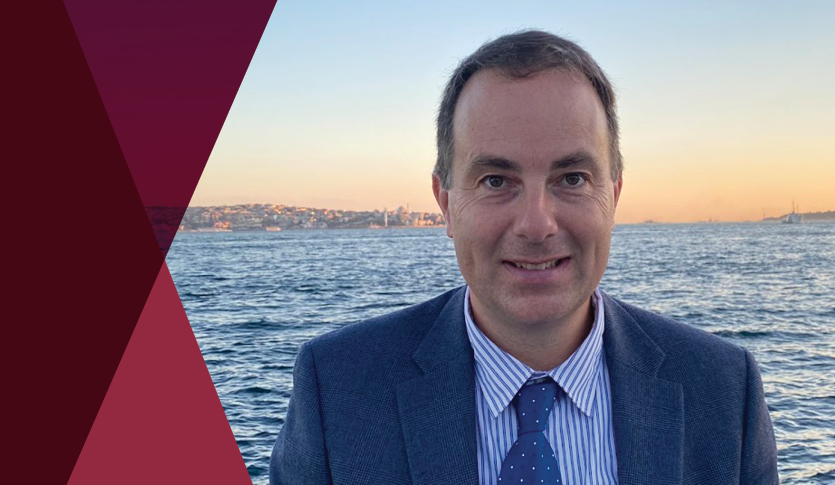 Douglas Cumming, professor of finance at Stevens Institute of Technology, wearing a blue suit and tie with a waterfront in the background. Maroon geometric shapes frame the left side of the image.