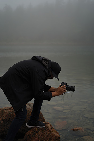 Vivek Kovvuru wears all black clothes and a hat and kneels on rocks in a body of still water as he takes a photograph with a professional camera.