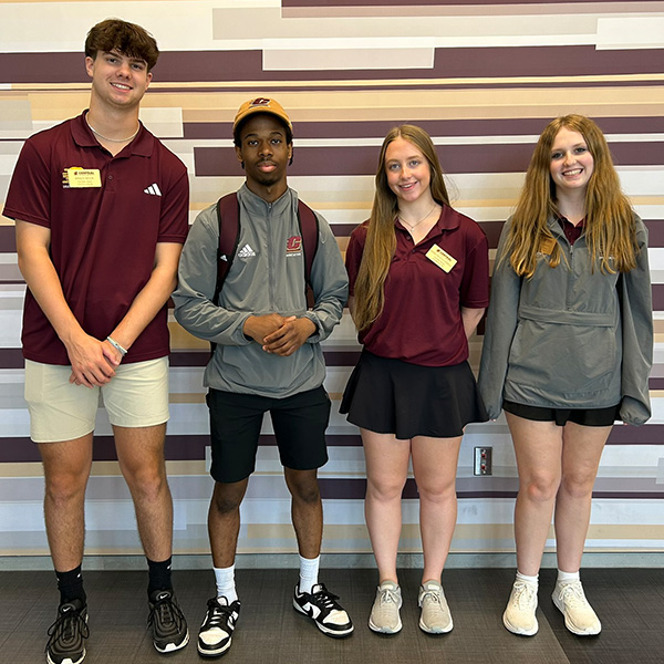 Four returning students acting as peer advisors stand in summer clothes, shorts and logo gear in CMU brand colors, wearing nametags in Grawn Hall. The students are arranged from tallest (two men) on the left to shortest on the right (two women).