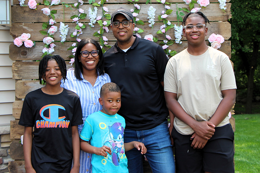 Jeremy Gibson wears a black hat, polo shirt and jeans, as he stands in front of a wooden wall with flowering vines and accompanied by his wife and three sons.