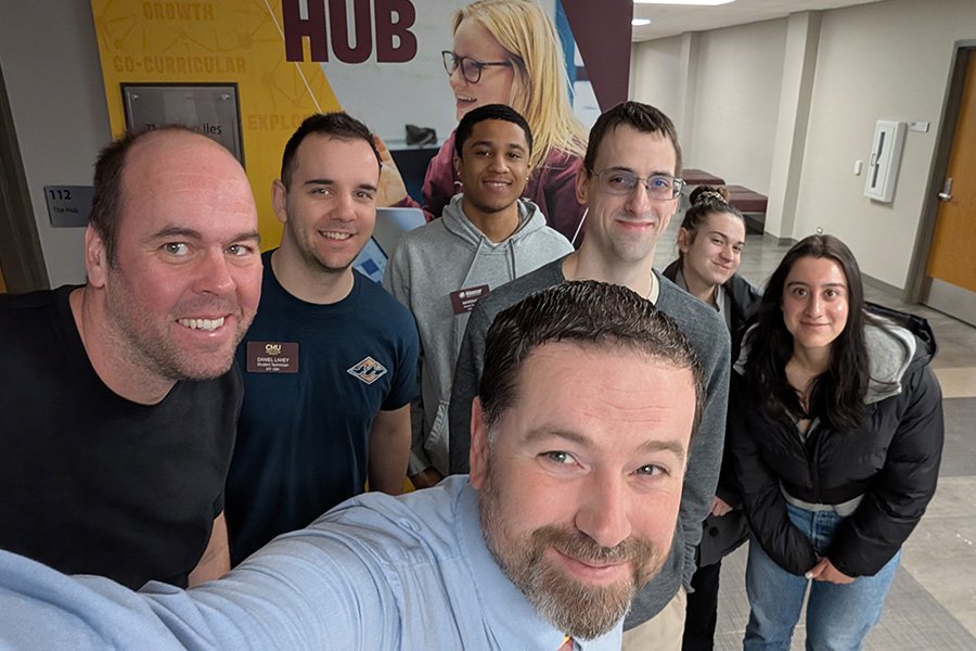 Tim Gramza holds his phone for a group selfie that includes his OIT team, including student workers. There are four men and two women behind him. They are standing in front of The HUB office.
