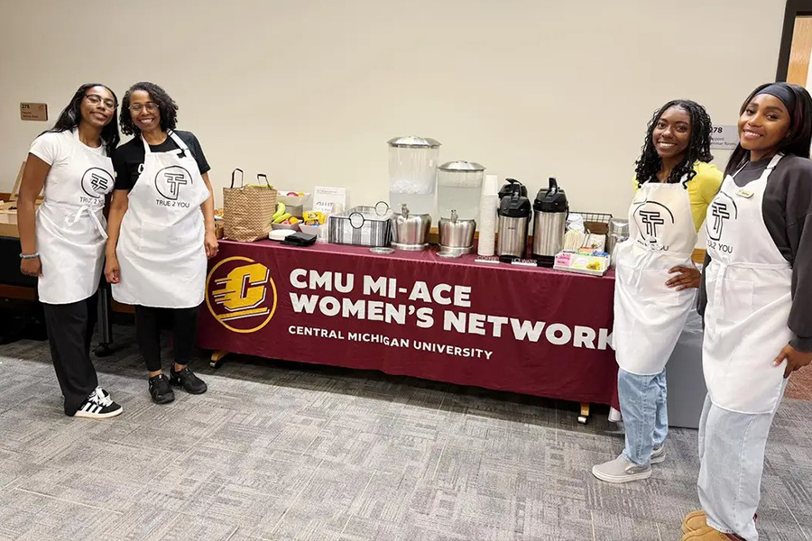 Four women from True2You candle making stand with aprons in front of a catering table with various breakfast items, coffee urns and water dispensers.