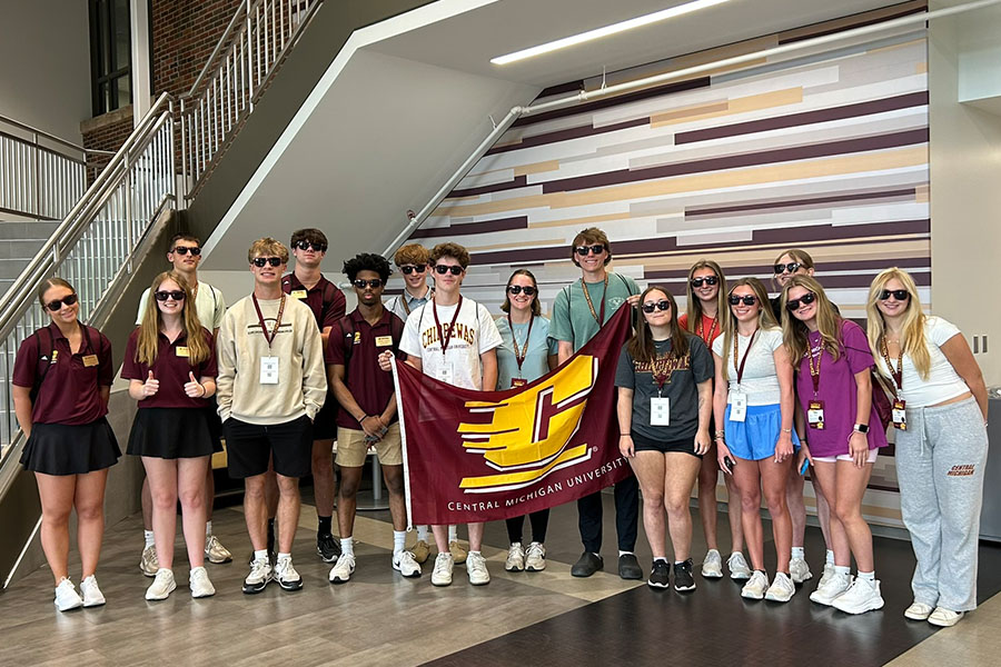 A group of incoming freshmen in summer clothes and matching maroon sunglasses stand with their peer advisors and hold an Action C maroon and gold flag in front of the Grawn Hall atrium stairs.