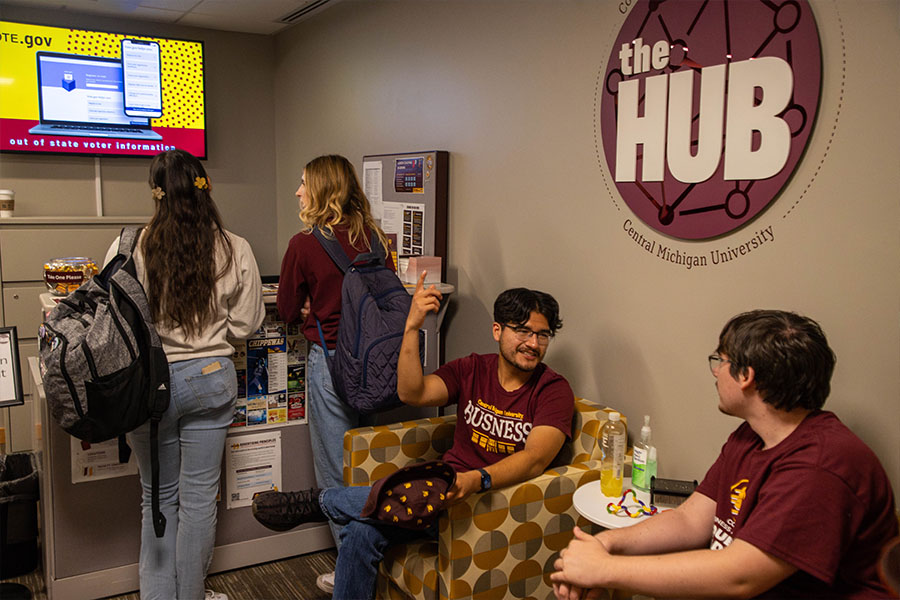 Two students sit in chairs while two additional students stand in front of a desk speaking to an Ambassador in the HUB.