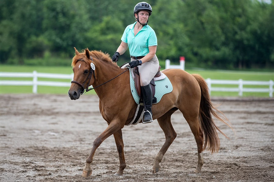 Alumna Stacy Salisbury wears a helmet and aqua short sleeve shirt while riding a brown horse in a pasture.