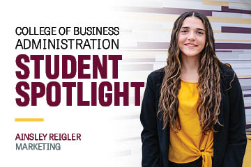 College of Business Student Spotlight focus marketing major Ainsley Reigler wears a black blazer over a gold shirt and poses at the top of the stairs in the Grawn atrium.
