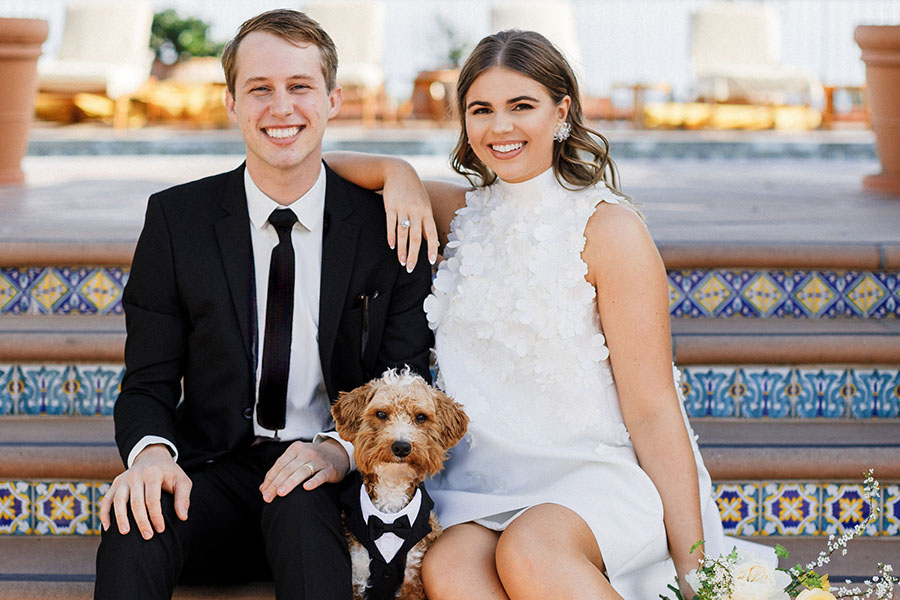 Tyler and Sarah Wallner are seated on a bench in their wedding clothes with their smally curly-haired dog (also in a tux) sitting between them.