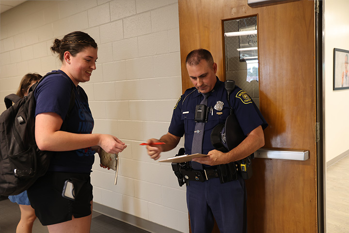A police officer checks to make sure a student is on the list near the door of an Accounting classroom. They are smiling as this is part of an introduction to Forensic Accounting at the College of Business.