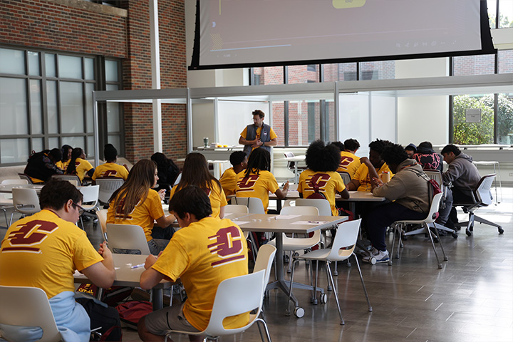 The HUB busy recruiting new students in the Atrium. There are many students seated in tables watching a presentation, all of them adorned in CMU gear.
