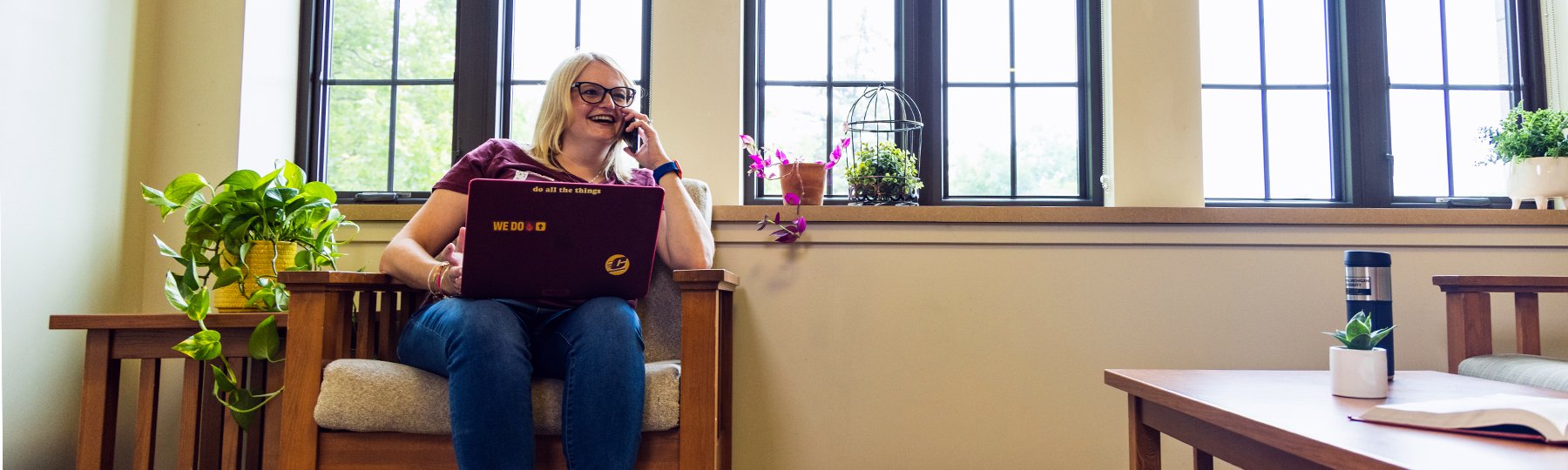 A smiling woman sits in a wooden chair by tall windows, talking on the phone with a maroon laptop on her lap. The laptop has stickers that say “WE DO” and “do all the things.” Plants and an open book decorate the cozy room.