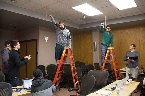 Lean Six Sigma workshop with students testing the flight of paper helicopters. Two students stand on laters as they drop the paper to the ground. Four other students observe with stop watches and record the time.