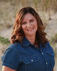 A professional headshot of Christine Witt wearing a denim, semi-casual button up blouse and standing in an outdoor location.