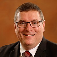 Professional headshot of Kevin McCarron in a black suit against a gray background.