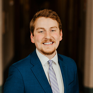 Owen Brinker smiles at the camera. He has short reddish brown hair and a goatee. He is wearing a blue suit jacker, white button down shirt and a silver striped tie.