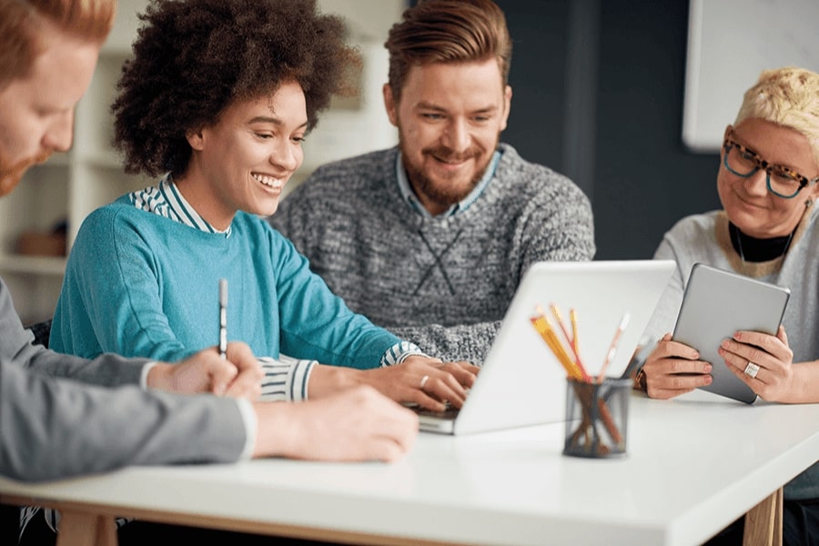 Four individuals are sitting at a desk, smiling and looking at a laptop.