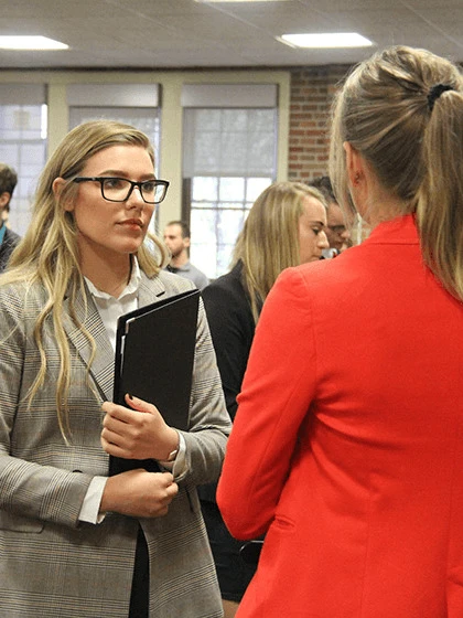 Two female students dressed in business attire standing and having a conversation.