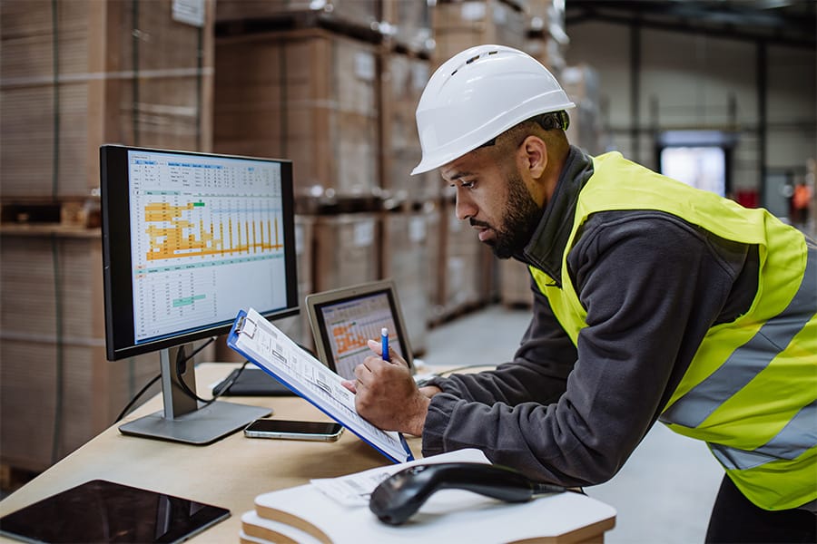 A man in a hard hat and hi-visibility vest looks at a clipboard while standing in front of a desk.