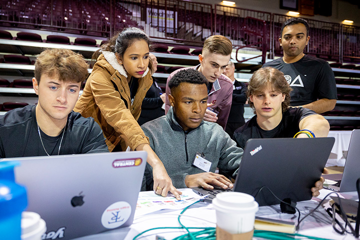 Students gather around laptops in McGuirk arena during the ERPsim. Six team members huddle over the screens intently, while a young woman points to information on a print out next to them. Cables and coffee cups are stacked around the laptops.