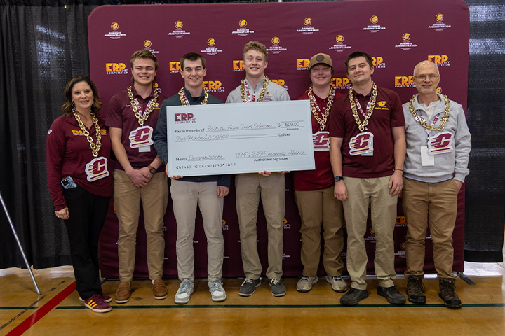 Students wearing large plastic chain necklaces with Action C logos post with faculty member Christine Witt and their ERPsim mentor. They hold a large first place check for winning an overall prize on their server.