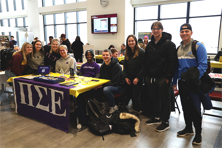 A diverse group of students are posing in front of a table advertising their Residential Student Organization. They are smiling and posing for the camera.