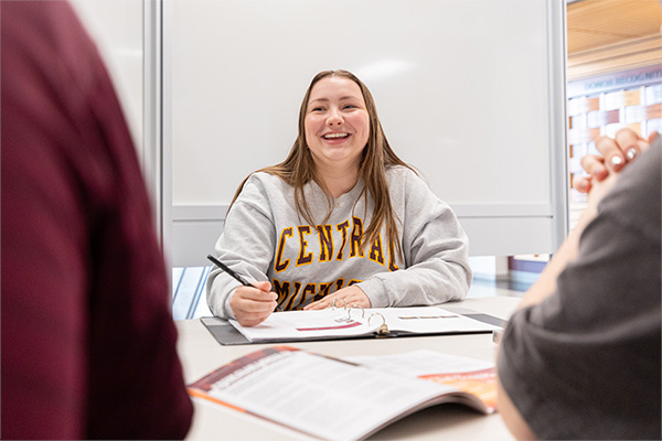 A female student is smiling as she works writes in a three-ringed binder. She's looking up towards another student who's unseen in the image. She's wearing a CMU sweatshirt and seated at a study space table on Grawn's first floor.