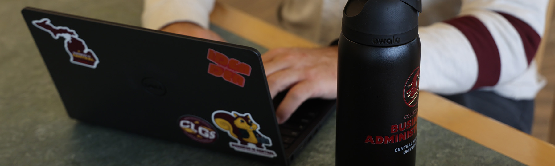 Hero-Cyber Sec 1800x540 A closeup picture of a person's hands working on a laptop on a table. The laptop has various CMU stickers on it, there's a CMU water bottle on the table as well.