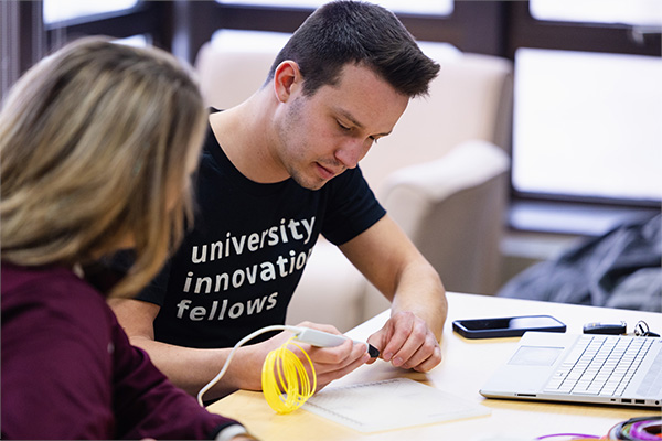 A student can be seen in consultation with a professor. They are seated at a table, the student is using a hand-held 3D printing device to create a prototype object.