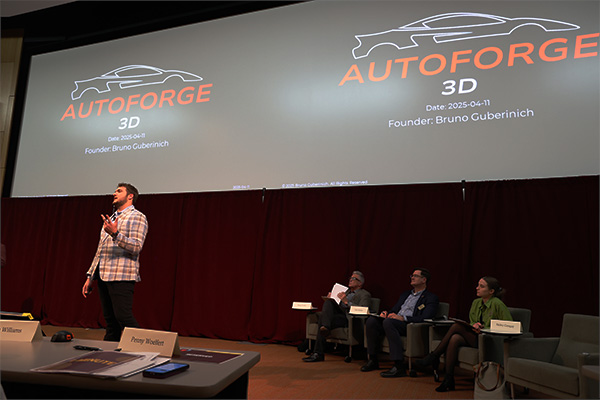 A student is giving a presentation at the New Venture Challenge. Their logo is prominently displayed on two screens behind them.