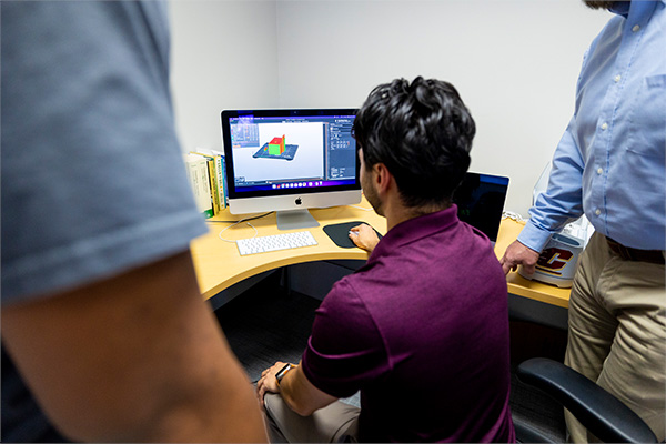 A student engineer is helping two people design a prototype in CAD. The image is from behind the group, a monitor is centered in the photograph, a design prototype can be seen on the monitor.