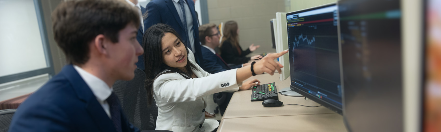 MFS 1800x540 Michigan Financial Scholars member, Carolina Hernandez Ruiz, shows her classmates how to use a Bloomberg lab terminal. She's talking to a male student and pointing at something on his monitor.