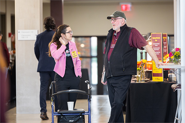 A student and a faculty member are in the hallway discussing her new venture idea. The student is listening intently, leaning on a wheelchair as her business venture idea is about handicap accessibility. The professor is standing near a table.