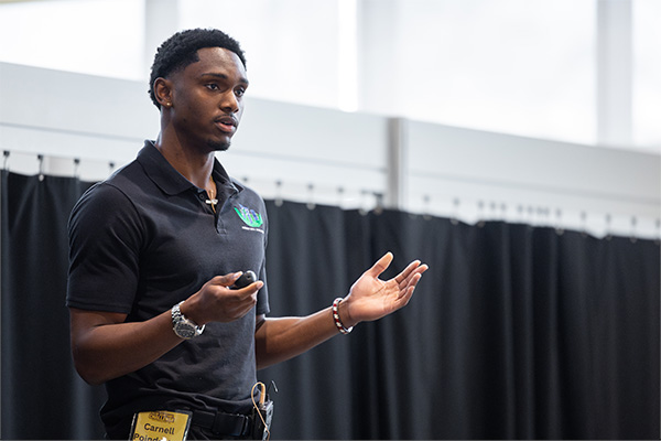 A student is on stage in Grawn Hall giving a presentation at the annual New Venture Challenge. The student is pitching for his educational outreach program.
