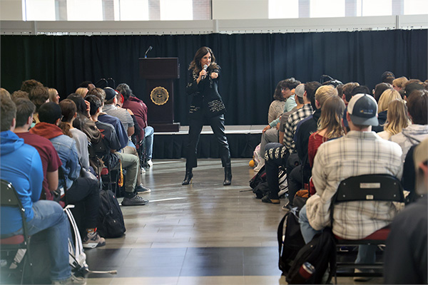 A keynote presenter is posing in front of a group of students during Grawn Hall's Dialogue Days. It's taken in action as she is giving her presentation. She's wearing a distinctive, custom suit with letters printed on it.